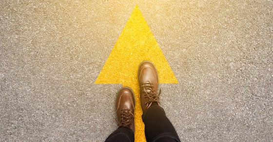 A top-down view of someone wearing black pants and brown shoes walking on a paved street with a large yellow arrow pointing in the direction they are walking