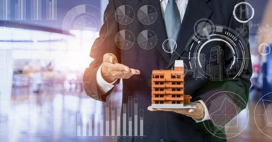 A person in business attire holding a small scale model of an apartment building while semi-transparent charts and graphs float in the foreground