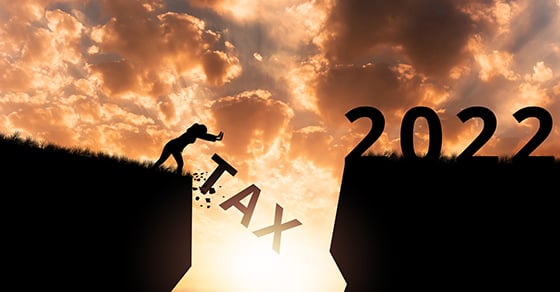 A person standing at the edge of a deep chasm while "2022" sits on the other side. The person is shoving the word "TAX" down into the chasm.