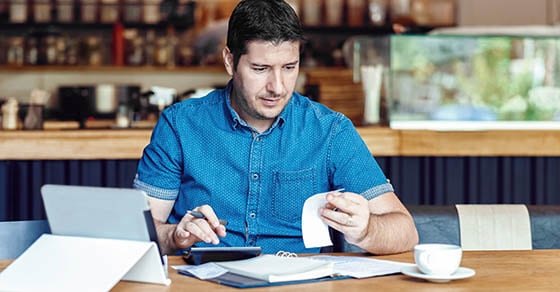 A male small business owner holding receipts and using a calculator.