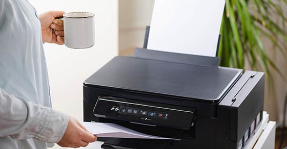 An office worker holding a coffee mug and pulling a document off of a printer.