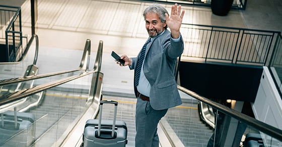 A man in a suit holding his phone while going down an escalator and looking back at the camera and waving. There is a suitcase next to him.
