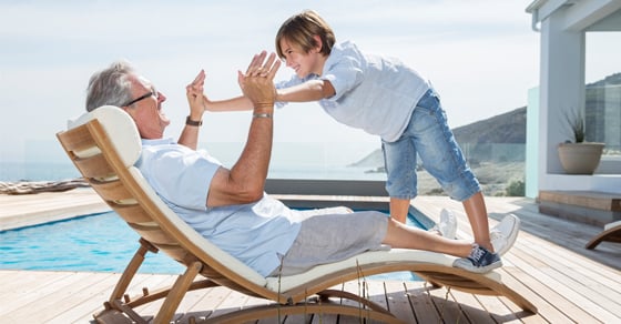 An older man sitting by a pool in a lounge chair holding his grandchild up with his arms while the child stands over him on top of the chair.