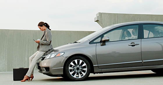 A woman in business attire leaning back on the hood of a car while looking at her phone.