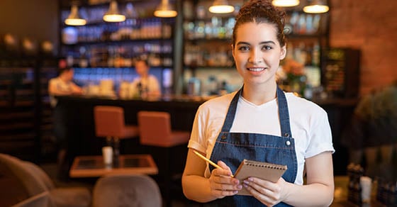A waitress standing in front of a bar in the restaurant holding a notepad and pen, as if she is ready to take a table's order.