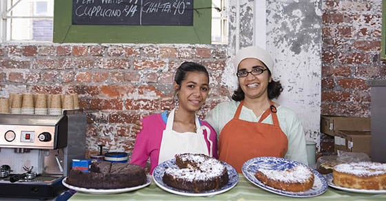 Two women smiling and standing behind a counter with cakes displayed on it.