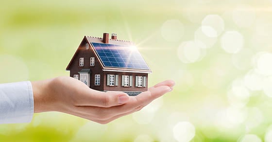 A hand holding a home with solar panels installed on the roof in front of a light green background.