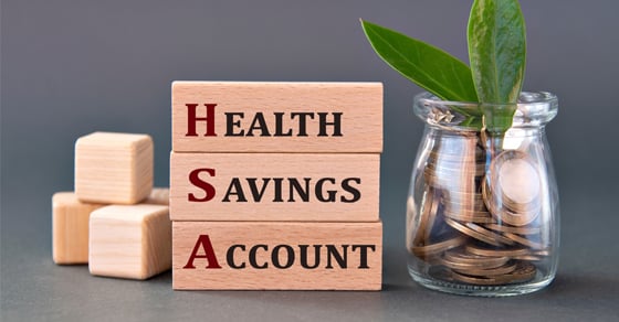 A gray surface that has stacked wooden blocks that read "Health Savings Account" and a glass jar filled with coins and a plant sitting on top of it.