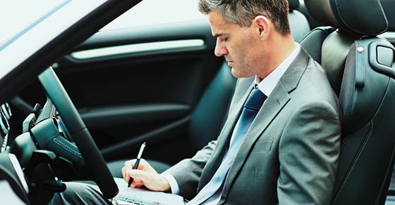 A business man sitting in a car while writing something on a notebook.