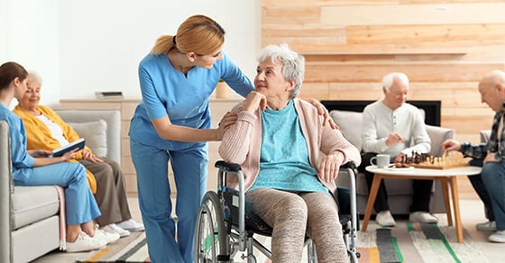 A nursing home employee speaking to a resident sitting in a wheelchair.