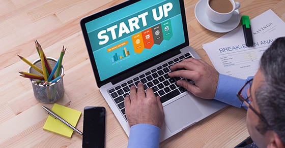 A man sitting at a desk looking at a laptop screen that reads "Start Up" with a graphic underneath.