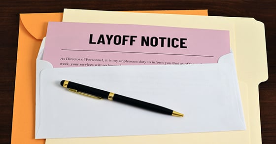 An envelope with a red piece of paper that reads 'LAYOFF NOTICE' sticking out of it, sitting on top of a couple different file folders.