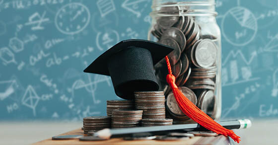 A lot of coins stacked on a desk and piled inside of a mason jar with a small graduation cap on top of them.