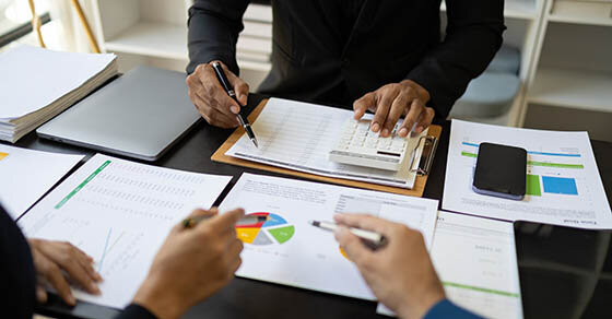 A view of a table with a lot of paperwork on it and three people standing around it discussing the paperwork.