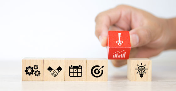 Five wooden blocks in a row with icons on them, and a hand placing a red block with a key icon on it into the row of blocks.