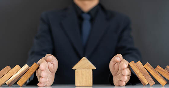 A house made of wooden blocks in the middle of a desk with a man sitting and blocking other wooden blocks from falling onto the house.