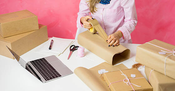 A person working at a desk that's cluttered with boxes, tape, scissors, and a laptop.