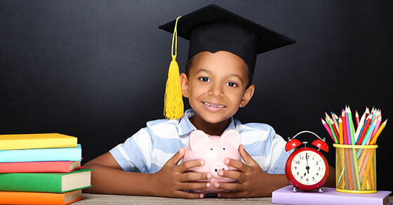 A child with a graduation cap on sitting at a desk holding a piggy bank.